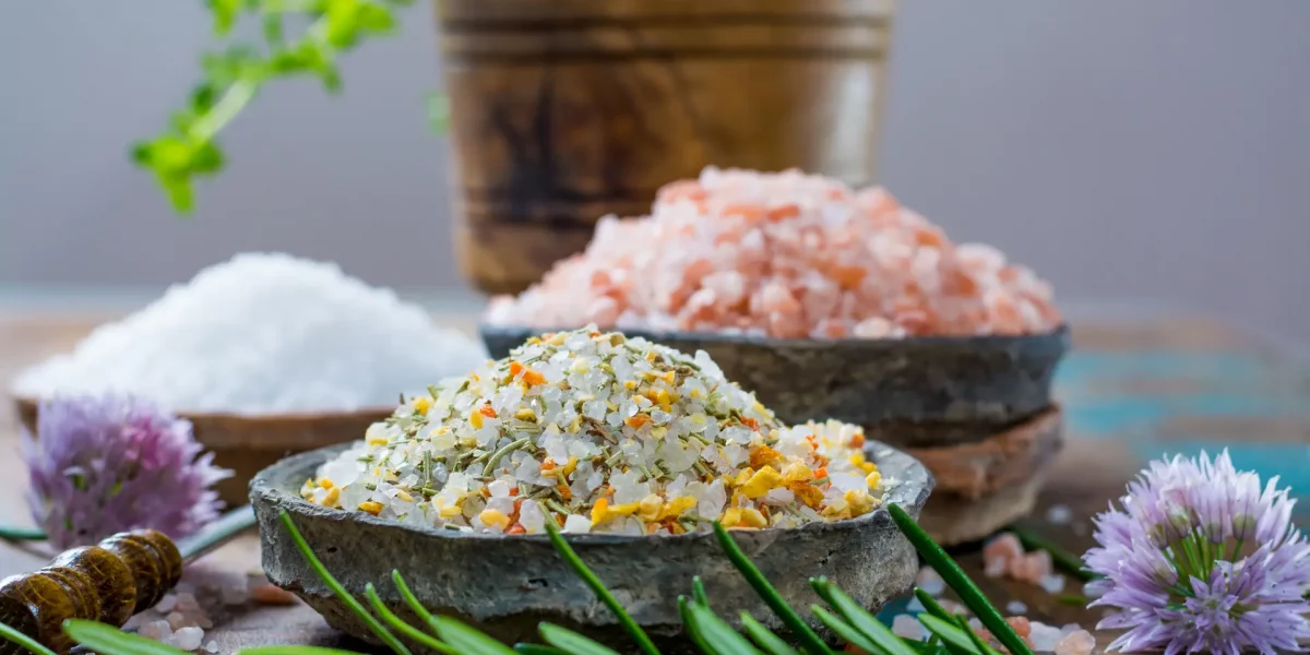 Different types of natural salt in stone bowls on wooden surface. White sea salt, pink Himalayan salt, Spiced salt with rosemary