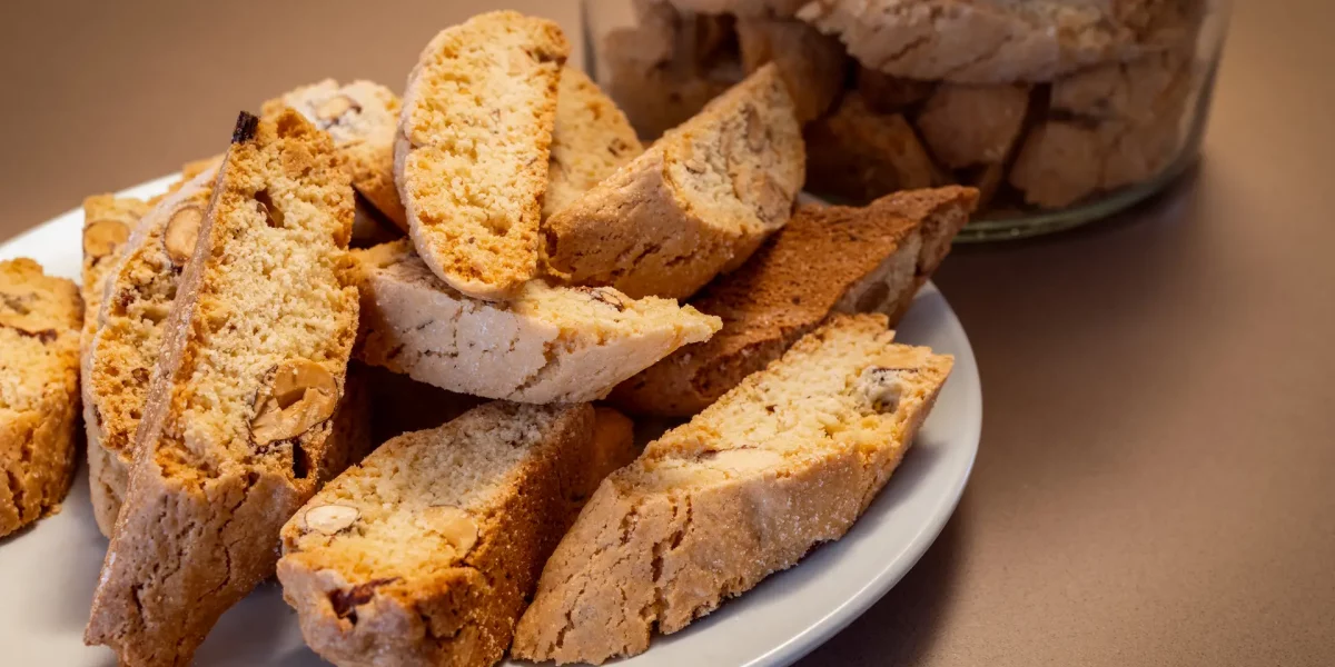 Several almond cantucci cookies rest on a plate with a jar of additional biscotti behind them The golden brown color and almond pieces show the crunchy texture of the cookies